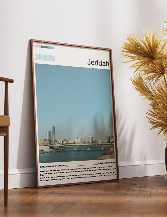 Framed poster of Jeddah on a wall next to a wooden chair and decorative plants.