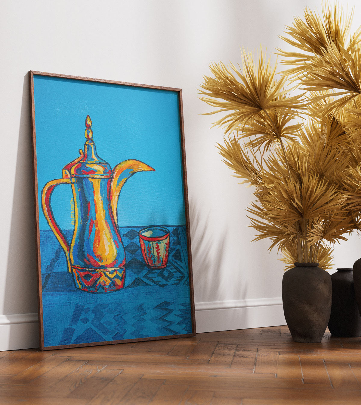 Framed artwork of a colorful teapot on a blue background, placed on a wooden floor with decorative plants.