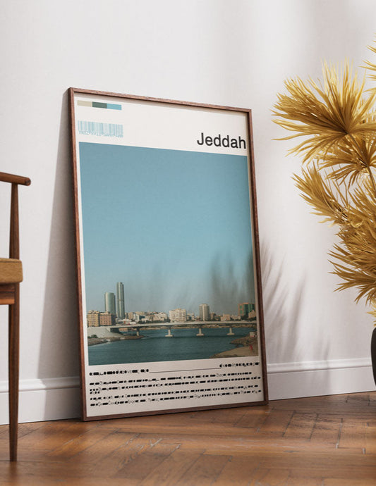 Framed poster of Jeddah on a wall next to a wooden chair and decorative plants.