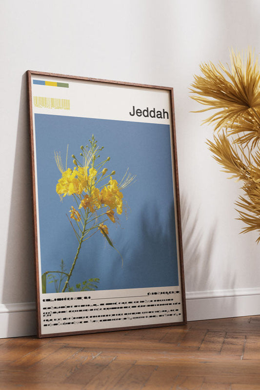 Framed artwork of a flower with 'Jeddah' text, placed on a wooden floor with decorative plants and a bench.